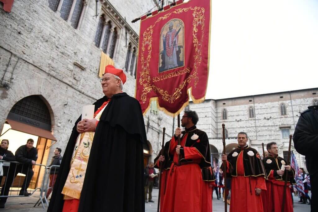 a-missa-é-a-mesma-em-todo-mundo (2) Cardeal em vestes litúrgicas vermelhas e pretas liderando procissão com bandeira religiosa em rua histórica de pedra.