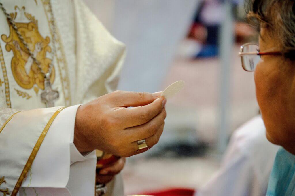Sacerdote entregando a Hóstia Consagrada a um fiel, simbolizando a Eucaristia como a fonte da Unidade da Missa.