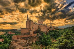 Vista panorâmica do majestoso Alcázar de Segóvia, um castelo medieval espanhol, com torres pontiagudas e muralhas de pedra robustas, sob um céu dramático e alaranjado de pôr do sol. Rodeado por uma paisagem verde exuberante, evoca a época da Inquisição Católica.