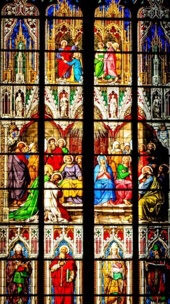 Vitral gótico detalhado e colorido dividido em registros horizontais. No topo, figuras divinas representam o Dogma; no centro, apóstolos e santos ilustram a Doutrina; na base, bispos e fiéis simbolizam a Disciplina e a vida da Igreja. A luz que atravessa o vidro une os três níveis em uma única estrutura de fé.