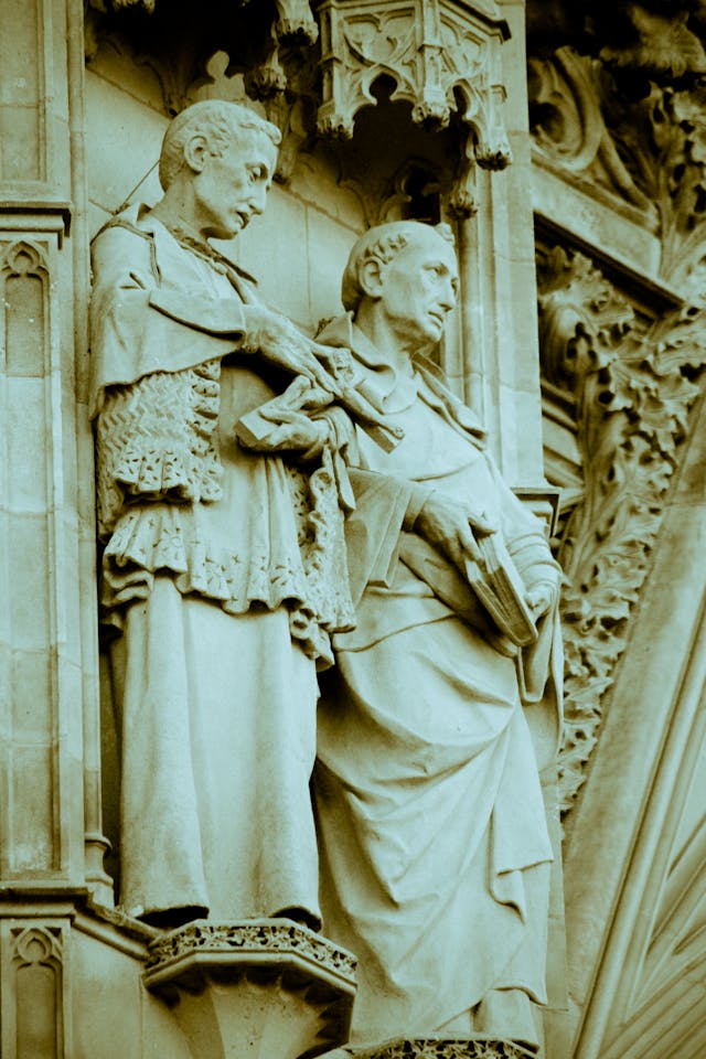 "Escultura em pedra de dois sucessores apostólicos em uma fachada de catedral. O gesto de um clérigo tocando o braço do outro simboliza a transmissão ininterrupta da fé e da autoridade, ilustrando a Tradição como uma corrente viva que remonta aos Apóstolos e não como uma criação humana posterior."